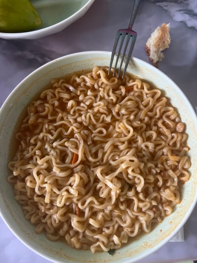 A close-up, overhead shot shows a bowl of instant ramen noodles with broth. The noodles are wavy and golden brown, mixed with small pieces of orange and green vegetables. A silver fork rests on the edge of the bowl, its tines submerged in the broth. To the right of the bowl, a small, torn piece of white bread with a browned crust lies on a patterned surface. In the background, out of focus, a portion of another bowl containing a green object, possibly a vegetable, is visible. The scene is brightly lit, suggesting an indoor setting, possibly a kitchen or dining area. The surface beneath the bowl appears to be a marble-patterned countertop. This image does not contain information about the city of Şamaldı-Say, Kyrgyzstan.