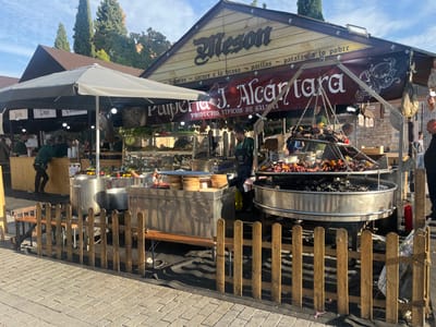 This image depicts an active outdoor food market scene, likely a festival or fair, located in Alcalá de Henares, Spain. The scene is set on a light-colored paved square, partially enclosed by a low, rustic wooden fence in the foreground.
Two primary food stalls are visible. The dominant stall on the right features a prominent wooden facade with a dark roof and a red banner. The banner reads "Meson" at the top, and "Pulpería J. Alcántara" below, with additional text indicating "PRODUCTOS TÍPICOS DE GALICIA" (Typical Products from Galicia) and offerings like "tripas," "carnes a la brasa" (grilled meats), "paellas," and "patatas a lo pobre." An octopus graphic is displayed on the banner.
This stall's focal point is a large, multi-tiered circular charcoal grill, approximately 1.5 to 2 meters in diameter, from which smoke ascends. Various cuts of meat, including sausages and larger pieces, are cooking on multiple levels suspended by chains above the coals. A male worker, dressed in a dark apron, is positioned behind this grill, attending to the food. To the far right, partially obscured, stands another male observer in a dark shirt. Cured hams or similar products are hanging from the stall structure to the right. A red fire extinguisher is visible on the ground beside the grill.
To the left, a second stall operates under a large, light-colored umbrella canopy and a darker rear canopy. This stall features a bamboo-paneled counter. Several individuals in green shirts are working behind this counter, preparing or serving. One person is leaning forward, attending to items at counter level. Large metal pots and various cooking or serving containers are visible on metal surfaces within this stall area. Shelves with produce or other items are also present.
The background reveals tall, slender coniferous trees and stone buildings consistent with historical architecture, all set against a clear blue sky with sparse white clouds. The overall lighting suggests bright daylight.