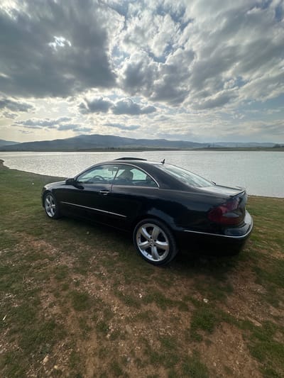 A black coupe, possibly a Mercedes-Benz CLK, is parked on grassy terrain near a body of water under a cloudy sky. The location is likely in or near Vrattsa, Bulgaria, indicated by landscape characteristics, although specific landmarks or signs identifying a street are not discernible. The car is angled towards the water. No individuals are visible in the image.
