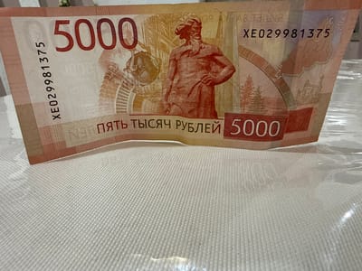 A close-up, slightly angled shot shows a 5000 Russian ruble banknote with the serial number XE029981375. The banknote is laid on a textured white surface, possibly a tablecloth, and is partially covered by a clear plastic sheet. The banknote displays a prominent image of a statue of Nikolai Rezanov, a Russian naval officer and statesman, on its left side, and a map of the city of Serebryakovka, Russia, on its right. The date "2023" is visible in the upper right corner of the banknote. The background is out of focus, but suggests an indoor setting with some ambient light.