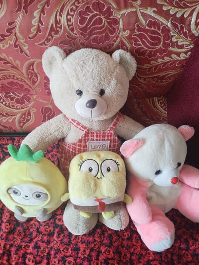 Four stuffed toys are arranged on a patterned surface. From left to right, they are a yellow sloth-like toy wearing a hood with a green stem on top, a yellow Spongebob-like character, a large beige teddy bear wearing red and white checkered overalls with a white patch on the bib that reads "LOVE", and a white teddy bear with pink ears and paws. The background is a red patterned fabric, possibly a couch or blanket. The lighting suggests it is daytime indoors.