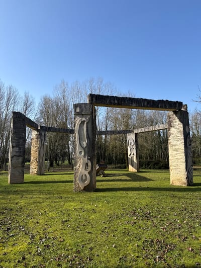 The image displays an outdoor stone sculpture complex situated on a green grass lawn in the city of Mealhada, Portugal. The installation features multiple large, rectangular pillars and horizontal lintels constructed from light-colored stone, forming an incomplete, rectilinear architectural framework. The structures exhibit varying surface textures; some pillars show vertical or horizontal grooving, while others display intricate, abstract carvings. A prominent central pillar features complex, organic-like designs, and a pillar positioned further back to the right reveals a stylized human-like figure. The ground consists of green grass with scattered brown leaves. In the background, a dense line of bare deciduous trees indicates a wooded area. The sky is clear and cloudless, displaying a bright blue hue. Strong sunlight illuminates the scene from the left, casting distinct shadows of the stone structures onto the grass. No individuals, animals, or specific actions are observed.