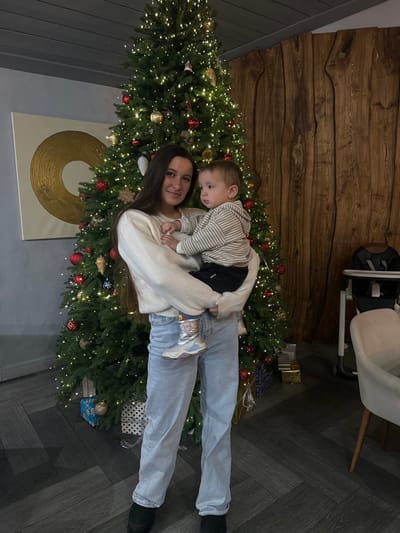 The image displays an indoor scene in Kalinovo, Russia, featuring a woman holding a child in front of a Christmas tree. The woman, estimated to be in her mid-20s to early 30s, possesses long dark hair and wears an off-white, ribbed long-sleeved sweater, light blue jeans, and dark footwear. She looks towards the viewer with a slight smile. She holds a young child, approximately 1-2 years old, dressed in a light-colored striped hooded top, dark pants, and metallic silver and pink boots. The child is looking towards the right of the frame.

The central background is dominated by a fully decorated Christmas tree, illuminated with warm white lights and adorned with red, gold, and silver baubles and other ornaments. At the base of the tree, several wrapped gifts in blue, gold, and white with black dots are visible. To the left, a gray wall features a large, framed abstract artwork depicting concentric gold circles on a white canvas. To the right, a wall composed of vertical, organic-shaped wooden panels is visible, alongside a white and black high chair and a partially visible light-colored dining chair with wooden legs. The floor is covered with dark gray material arranged in a herringbone pattern.