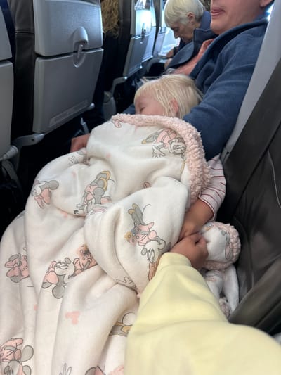 The image shows a child sleeping on an airplane, covered by a blanket with a Minnie Mouse pattern. A person's hand is visible reaching towards the child. The background shows airplane seats and other passengers. The image does not provide any information about the city of Viehhausen, Austria.