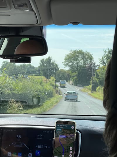 The image captures the interior of a car driving through a rural area, likely in or around Derrylin, United Kingdom, given the navigation displayed on the phone. The view is through the windshield, showing a two-lane road with greenery and trees lining the sides. Two vehicles are visible on the road ahead: a silver car in the immediate foreground and a yellow bus further up the road. The car's dashboard is visible, featuring a digital display showing the current speed as 66 km/h and the date as 2026-07-19. A smartphone, mounted on a holder, displays a navigation app showing a route on "Belturbet Rd, A509." A rear-view mirror is also visible, reflecting the head of a person. The lighting suggests it is daytime with partly cloudy skies.