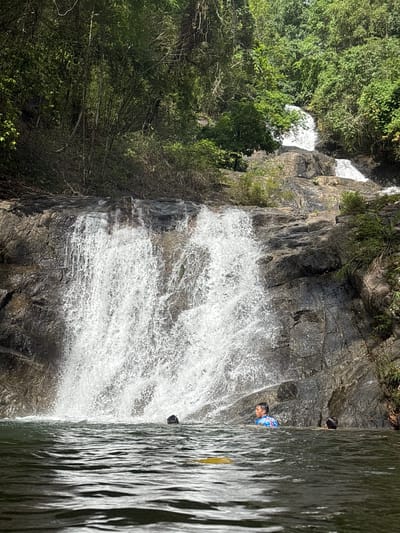 A waterfall cascades down a rocky cliff in Thai Mueang, Thailand. The surrounding area is lush with green trees and vegetation. Two individuals are visible near the waterfall; one person in a blue and white shirt is partially submerged in the water near the falling water. The other individual, above them, is further from the waterfall.