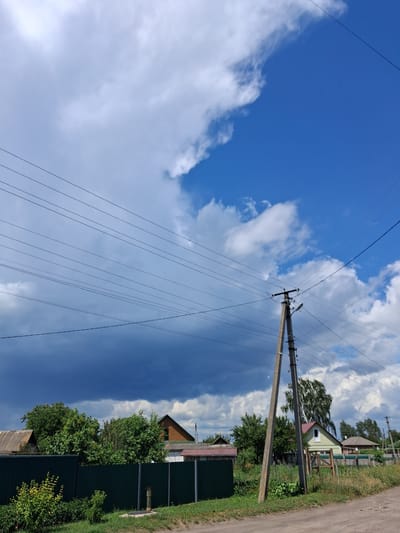 The image depicts a rural landscape with a backdrop of a dramatic sky. Overhead, numerous power lines crisscross the frame, strung between a tall concrete utility pole on the right and an unseen point to the left. The sky is a mix of bright blue and dark, ominous clouds, suggesting an approaching storm or recent rain.
In the foreground, a dark green metal fence runs horizontally across the image, separating the road from a residential area. Beyond the fence, several houses and trees are visible. The houses appear to be modest, with pitched roofs. Dense green foliage from trees and bushes fills the space between the houses and obscures parts of the structures. The ground in front of the fence is covered in green grass, with a dirt road running along the bottom edge of the frame. A yellow, metallic object, possibly a well or a pump, is visible near the fence line.
The scene is devoid of any people or animals. The overall impression is one of a quiet, semi-developed rural area under a dynamic sky. The location is identified as Krolevets, Ukraine, suggesting a setting in that specific region.