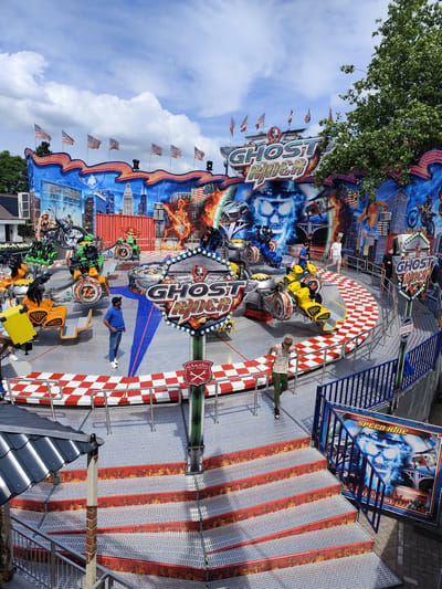 The image depicts a bustling amusement park ride called "Ghost Rider" in Best, Netherlands. The ride features brightly colored vehicles resembling motorcycles and cars, set against a backdrop of cityscapes, flames, and American flags. Several people are visible, some riding the attraction, others observing or walking nearby. The ride itself is elaborate, with flashing lights, a checkered floor pattern, and promotional signage. The overall scene suggests a lively and exciting atmosphere at the fairgrounds.