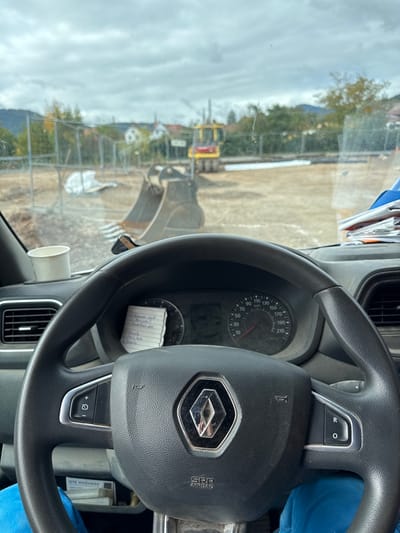 The image presents a first-person view from inside a stationary Renault vehicle, looking forward through a dusty windshield at an active construction site in Scherwiller, France, under an overcast sky.

In the immediate foreground, a black Renault steering wheel with a silver diamond logo and "SRP AIRBAG" inscription is centrally positioned. Control buttons are present on both the left and right spokes of the steering wheel. Behind it, a crumpled piece of white paper with handwritten text is tucked into the dashboard, partially obscuring the instrument panel, which includes a speedometer with markings up to 210 km/h. To the left on the dashboard, a white disposable cup is visible. To the right, a stack of papers and folders rests on the dashboard. The lower portion of a person's legs, clad in blue pants, is visible at the very bottom of the frame, indicating a driver or passenger.

Beyond the vehicle's interior, a large, dark-grey excavator bucket occupies the midground, positioned on cleared, light-brown earth. Further back on the site, a yellow road roller (compactor) is visible. A temporary fence, possibly chain-link, delineates parts of the construction area, behind which white tarpaulins or construction materials are present. In the background, residential buildings with traditional roofs are visible amidst trees displaying some autumn foliage, indicating proximity to a populated area. Distant hills or mountains frame the horizon beneath a uniform grey, cloudy sky.