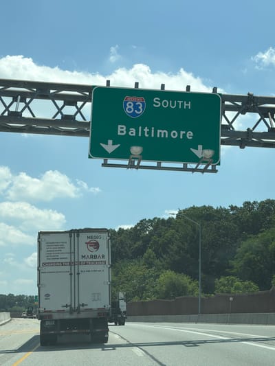 In York Township, United States of America, a green highway sign indicates "INTERSTATE 83 SOUTH BALTIMORE" with downward pointing arrows for both lanes. Below the sign, a white semi-trailer truck with the logo "MARBAR" is traveling on the highway. The truck has text on its side detailing lease and purchase programs for trucks. The scene is under a bright, partly cloudy sky, with lush green trees lining the side of the highway.