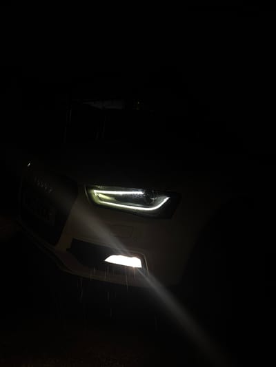 A white Audi car is parked in the dark, possibly in Dolni Tsibar, Bulgaria. The car's headlights are on, illuminating the road and what appears to be rain or water droplets in the air. The license plate is partially visible and reads "VN2 RRK". The image focuses on the front of the car, with the grill and one headlight prominently featured. The background is completely dark, suggesting it is nighttime. There are no people visible in the image.