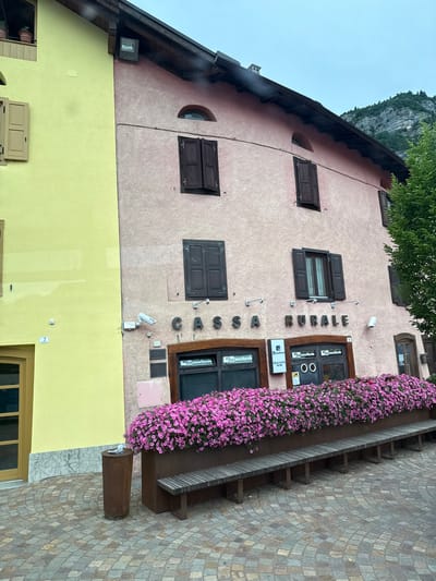 The image shows the exterior of a building in Fai della Paganella, Italy. The building has a pink facade with several windows and shutters. Above the entrance, large metal letters spell out "CASSA RURALE." Below this, there is a storefront with a window displaying "La Cassa Rurale Sportello ATM." A long planter filled with vibrant pink flowers runs along the front of the building, with a wooden bench placed in front of it. A cylindrical trash can is located to the left of the planter. The pavement in front of the building is paved with cobblestones arranged in a circular pattern. A green tree is visible on the right side of the image, and a mountain is in the background. The sky is overcast.