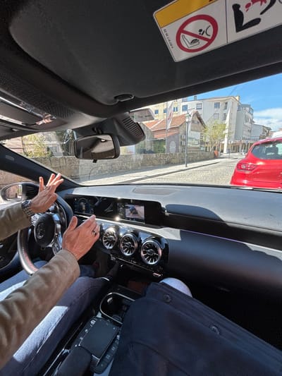 The image depicts a forensic view from inside a vehicle looking forward onto a cobblestone street in Chaves, Portugal. The car's interior features a black dashboard with three prominent circular air vents, a digital instrument cluster, and a central infotainment screen displaying a vehicle camera feed. A black multi-function steering wheel with a Mercedes-Benz emblem is partially visible on the left. A "Via Verde" transponder is affixed to the windshield above the rearview mirror. A sun visor on the upper right displays an airbag warning decal.
The driver's forearms and hands are visible, gesturing with open palms above the steering wheel. The driver wears a silver-toned wristwatch on the left wrist and a dark ring on the right ring finger. The driver's attire includes a light brown long-sleeved garment and blue jeans. The legs of a front passenger, clad in dark trousers and light-colored shoes, are visible in the lower right of the frame.
The exterior scene reveals a cobblestone street. A red five-door hatchback is positioned ahead, facing away from the viewer, stopped or parked. To the left, a substantial stone wall supports buildings typical of urban Portuguese architecture. A light-colored two-story building with a red-tiled roof is visible behind the wall, featuring a large mural. A red and white flag is partially visible near this structure. In the background, under a clear blue sky, larger multi-story buildings with light-colored facades and numerous windows define the urban landscape of Chaves. The scene is illuminated by bright daylight.