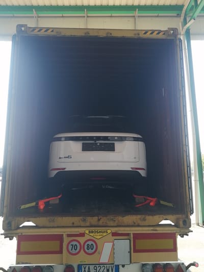 The image shows a white Range Rover SUV being loaded into a shipping container. The container is yellow and appears to be on a trailer. The vehicle is positioned facing forward within the container. There are orange straps and yellow tie-downs visible, suggesting the car is secured for transport. The background shows the open doors of the container and the underside of a roof structure, possibly part of a warehouse or loading dock. On the trailer, there are regulatory speed limit signs (70 and 80), hazard markings, and license plates. A sign on the trailer reads "BROSHUIS HOLLAND". The scene is located in the city of Borgolavezzaro, Italy, as indicated by the license plate which begins with "XA" and an indication of the location within the frame of the image.
