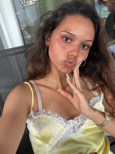 A young woman with long, dark hair is lying down, taking a selfie. She is wearing a light yellow slip with white lace trim. Her eyes are open and she is looking at the camera, with her lips pursed. Her right hand is touching her cheek, and she has long, manicured fingernails. She is also wearing a gold watch on her left wrist. The scene appears to be outdoors, possibly on a patio, with a textured dark surface beneath her. In the background, through what looks like a window or glass door, a reflection shows a partially obscured view of plants and possibly furniture. The lighting suggests it is daytime.