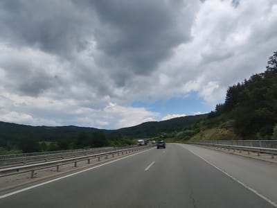 The image captures a view from a vehicle traveling on a highway, likely in or near Golemo Buchino, Bulgaria. The highway curves to the right, with a metal guardrail lining the left side. The road is asphalt, with a white dashed line marking the lanes. In the distance, on the left side of the highway, several large trucks are visible, suggesting a traffic slowdown or congestion. A black SUV is in the foreground, on the right lane of the highway, moving away from the camera. The surrounding environment is hilly and densely forested with various shades of green. The sky above is a mix of dark, heavy clouds and patches of bright blue sky with white clouds, indicating an overcast or partly cloudy day. The lighting suggests it is daytime.