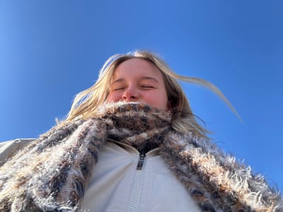 A low-angle portrait of an individual, primarily showing their upper body from the chest up, set against a clear blue sky. The subject, a person with blonde hair, is wearing a light-colored jacket over which a multi-colored (brown, black, white, beige) fluffy scarf is wrapped, concealing the lower portion of their face. Their eyes are closed, and a subtle smile is evident. A small metallic ring is present in the left nostril. Strands of their blonde hair are visibly swept to the right, indicative of wind. The background is a uniform, bright blue sky with no clouds. The precise location within Kingston, Canada, cannot be identified from the visual data within the image.