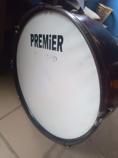 The media file displays a close-up, angled shot of a large drum, likely a bass drum, positioned indoors in Warri South, Nigeria. The drum is the central subject, featuring a prominent white drumhead with the brand name "PREMIER" printed boldly in black capital letters. Below "PREMIER," there are faint, faded, circular markings and other indistinct text or a logo that is not clearly legible.

The drum's shell appears to be a dark, possibly reddish-brown or burgundy color, and it is fitted with chrome or silver-colored tension rods and lugs around its circumference. The drumhead shows some signs of wear, including minor scratches and scuffs. The drum is resting on a light-colored tiled floor, which is visible in the lower part of the image.

In the blurred background, hints of other objects can be seen, including what appears to be a woven basket or container on the right, and possibly some blue fabric or another object. There is no specific activity taking place; the drum is stationary. The lighting suggests an indoor environment, possibly with indirect natural light or artificial light. No people or specific events are depicted.