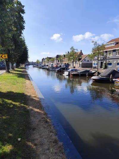 The image depicts a canal in the city of Sneek, Netherlands. The canal runs through a built-up area with buildings lining one side and a tree-lined grassy bank on the other. Several boats are moored along the canal. The sky is blue with some scattered clouds. A yellow sign with a black number "4" is visible on the left side of the image. The buildings appear to be a mix of residential and commercial properties. The water in the canal is calm and reflects the sky and surrounding structures. The scene suggests a pleasant day in a Dutch city.