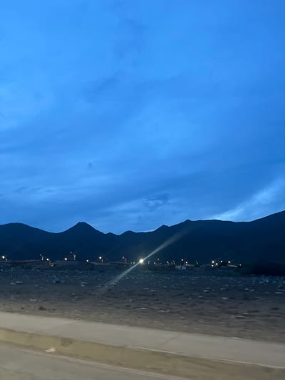 The image depicts a nocturnal or twilight scene in a semi-arid landscape, likely on the outskirts of San Antonio, Venezuela. The sky is a deep, moody blue with wispy clouds, suggesting the end of daylight. In the background, the silhouettes of mountains are clearly visible, their rugged outlines softened by the low light. Along the base of the mountains, a line of streetlights illuminates a developing settlement or small town. Buildings, some with terracotta-colored roofs, are discernible, and the lights create a scattered pattern across the landscape. The foreground consists of a dry, rocky terrain, with a paved road or pathway running through it, the surface of which is partially visible and appears to be a pale color. The overall impression is one of a remote, developing area under a vast, darkening sky.