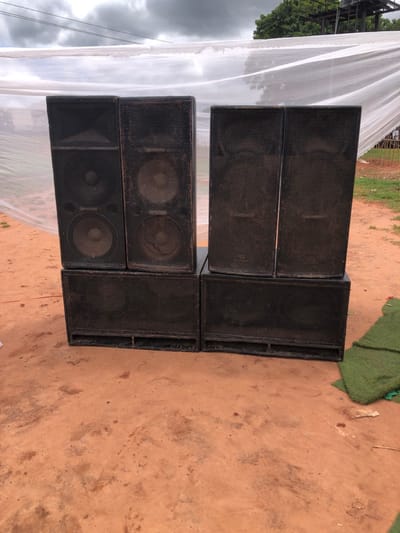 The image displays an outdoor scene in Amanuke, Nigeria, featuring professional audio equipment. Six black speaker cabinets are arranged in two stacks on reddish-brown, compacted dirt. Each stack consists of two taller, narrower speakers placed atop a single wider, shorter subwoofer cabinet. All speakers have metal grilles covering their cones and show signs of dust and use. In the background, a translucent white fabric, possibly part of a canopy or tent, stretches across the upper portion of the frame. Beyond this fabric, green foliage from trees and a dark, multi-level metal structure are visible against an overcast sky. To the right of the speaker stacks, a piece of green artificial turf is folded or rolled on the ground. No individuals or active interactions are present in the scene.