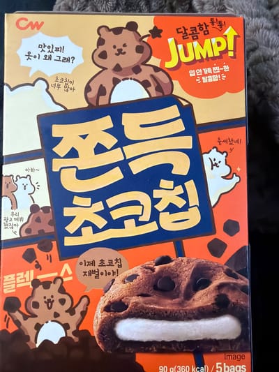 The image displays a close-up view of a food product box with Korean text and cartoon graphics. The box is predominantly orange and yellow, with a large blue panel featuring large yellow text. The main graphic on the box is a cartoon of a chocolate cookie sandwich with white filling and chocolate chips. There are also illustrations of cartoon bears on the box, some of which are speaking in speech bubbles. The text on the box is primarily in Korean, with some English words like "JUMP!". The bottom right corner of the image shows "Image 90 g(360 kcal) / 5 bags". The image is sharp and well-lit, with the product box being the central focus. There is no indication of any location in Tofta, Sweden, or any environmental context outside of the product packaging itself.