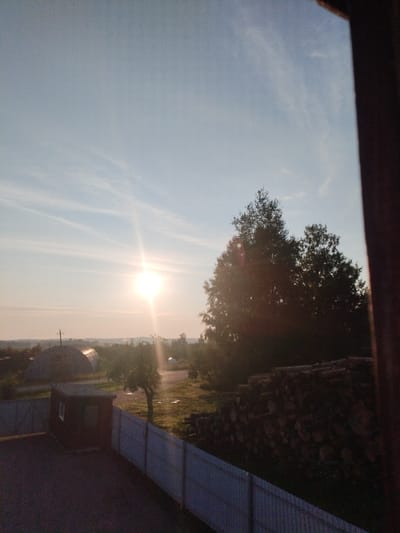 The image depicts a rural scene at sunset, likely within the Вароніцкі сельскі Савет region of Belarus. The sun is low in the sky, casting a bright glare and long shadows. In the foreground, a dark, asphalted area is bordered by a light-colored metal fence. To the left of the fence is a small, dark red shed with a single window. Further back, a larger area is visible with scattered trees and some structures in the distance, including what appears to be a rounded, agricultural building. On the right side of the image, a large pile of cut logs is stacked. A large, dark green tree dominates the middle right portion of the frame, with branches partially obscuring the sky. The sky itself is a pale blue with thin, wispy clouds. A dark brown, vertical element, possibly a doorframe or window frame, is visible along the far right edge of the image.