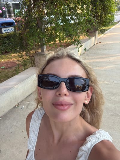 A young woman with blonde hair and fair skin is taking a selfie outdoors. She is wearing black sunglasses and a white, ruffled top. Her lips are slightly parted, and she appears to be smiling. A star-shaped earring is visible on her right ear. The background shows a concrete path next to some greenery and trees. In the distance, a light blue vehicle with some signage is visible, partially obscured by the foliage. The lighting suggests it is daytime. The image is oriented vertically, with the woman's face taking up a significant portion of the frame. There is no visible text in the image.