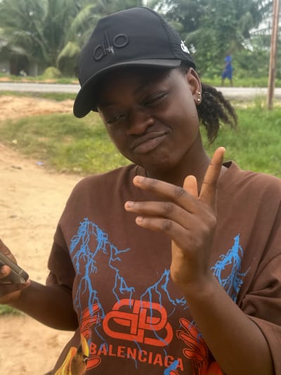 A young Black woman, wearing a black baseball cap with an "alo" logo and a brown t-shirt with a blue lightning bolt design and a red "BB" logo, poses outdoors in Ganyingbo, Nigeria. She is winking and holding up two fingers in a peace sign with her right hand. Her left hand is holding a smartphone. The background consists of greenery, dirt, and a distant figure walking on a road.
