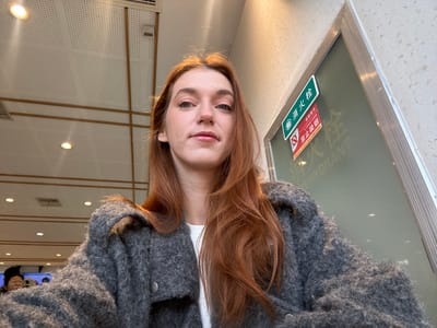 A woman with red hair and fair skin looks directly at the camera. She is wearing a gray, fuzzy cardigan over a white shirt. Behind her is a room with a white, perforated wall and recessed lighting. A window with signs and writing is partially visible on the left. To the right, a person is reflected in a screen. The photograph is rotated 180 degrees.