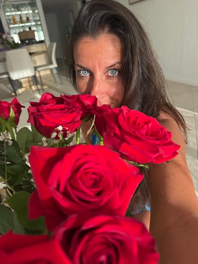 A close-up, self-portrait photograph depicts an adult female positioned in a residential interior in Lely, United States of America. The woman has dark brown hair and blue eyes, which are directed at the camera. Her face is partially obscured by a bouquet of approximately five to seven red roses, interspersed with green foliage and small white flowers, held in the foreground. Her right arm and shoulder are visible on the right side of the frame. The background shows a modern living or dining area featuring a light-colored dining table with two chairs to the left, and a white cabinet with glass doors containing bottles further back. The floor appears to be a light-colored tile or polished surface. The scene is brightly illuminated by interior lighting.