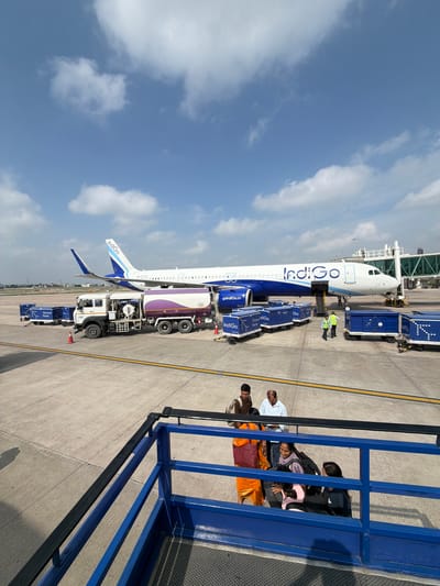 An Indigo Airlines airplane is parked at an airport tarmac in Vikram Nagar, India, under a partly cloudy sky. The plane, with the airline's blue and white livery, is connected to a jet bridge on the right side of the frame. In front of the aircraft, a white fuel truck with purple accents is parked next to several blue baggage carts, some of which are labeled "Indigo" and "Ground Force." Two airport workers in high-visibility vests are near the baggage carts. In the foreground, passengers, including women in traditional Indian attire and carrying luggage, are visible near a blue ramp structure. The ground is paved concrete, with yellow and white markings.
