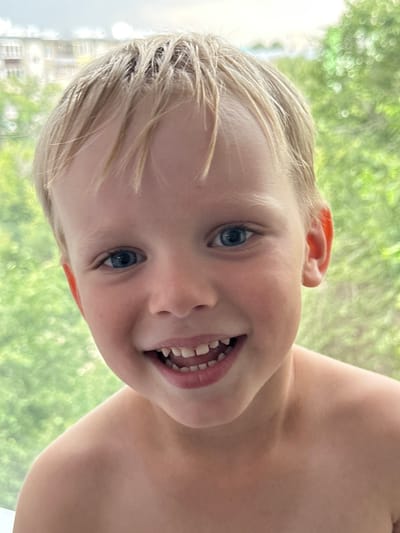 A close-up, vertical, eye-level shot captures a young boy with blond hair and blue eyes smiling at the camera. He is bare-chested, and his shoulders are visible. The background is out of focus, showing a blurred expanse of green foliage and a building. The image was taken outdoors, likely on a bright, overcast day.