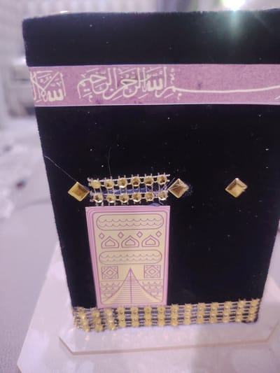 The image shows a close-up of a black, velvet-covered object, possibly a decorative item or a small model, resembling the Kaaba. It is adorned with gold-colored embellishments. A band of light purple material with Arabic calligraphy in white is wrapped around the top of the object. Below this band, there are rows of small, gold, diamond-shaped gems and a patterned section with wavy lines and geometric shapes, including what appears to be a pyramid. The object is placed on a white surface, which could be a display stand. There is no indication of the city of Bahshitpa, Uzbekistan, or any specific location within it. The image focuses solely on the object and its decorative elements.