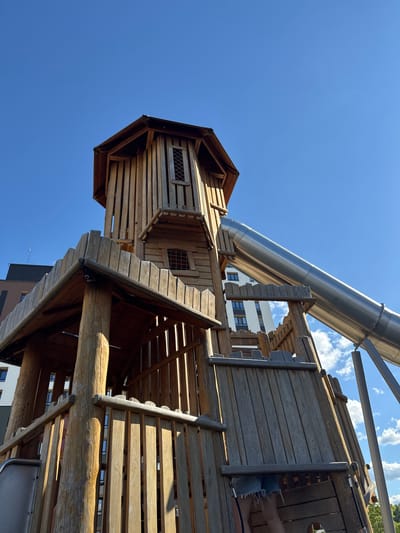 This image features a wooden playground structure with a large, silver slide. The structure appears to be a multi-level playhouse or fort, built with logs and wooden planks, some of which show signs of wear. A silver tube slide angles downwards from a higher platform. In the background, a modern white building with dark windows is visible, suggesting an urban or semi-urban setting. The sky is bright blue with scattered white clouds, indicating it is daytime and likely sunny. The perspective is from below, looking up at the play structure and the sky. There is no visible text in the image. The scene is set in Patrusheva, Russia.