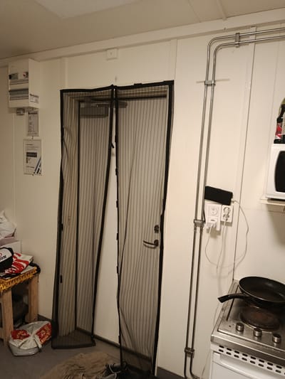 The image shows an interior scene, possibly a utility room or small kitchen, within a building in Vallberga, Sweden. The room is sparsely furnished and appears somewhat utilitarian.
In the center of the image, a double door is covered by black magnetic screen doors, which are partially open, revealing a white door behind. The screen doors have vertical stripes and appear to be made of mesh material.
To the left of the doors, a white electrical panel with multiple switches and indicator lights is mounted on the wall. Below the panel, there are two framed documents attached to the wall. A small wooden shelf unit with some items on it, including a red and white hat or cap, is positioned to the left of the screen doors. A gray plastic bag with red markings is on the floor near the shelf.
To the right of the doors, a white wall features several electrical outlets, one of which has a black smartphone plugged in and charging. Metal conduit runs vertically along the wall. A microwave oven is mounted on a shelf to the right, with a black cooking pan placed on a stovetop below it. The stovetop is part of a white kitchen appliance, likely a stove or range.
The walls are painted white, and the floor appears to be a dark gray linoleum or concrete. The lighting in the room is artificial and somewhat dim. There are no people visible in the scene. The overall impression is of a functional but not particularly well-maintained space.