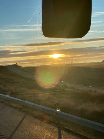 A horizontal view through a vehicle windshield captures a sunrise or sunset. Streaky clouds fill a blue sky on the left, contrasting with a bright sun over a distant horizon. Reflections and glare obscure details, with a large lens flare dominating the right side. A portion of the vehicle's interior, including a rearview mirror, is visible on the left. A highway guardrail and the road surface are seen to the right of the windshield.