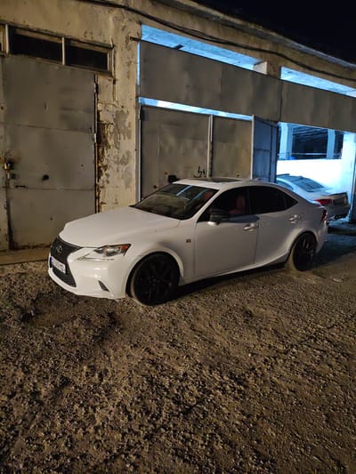 A white Lexus IS sedan is parked on a gravel surface in front of a large, weathered garage with metal doors. The car is angled towards the right, with its front facing slightly left. The garage structure shows signs of age, with peeling paint and concrete. Bright blue light emanates from within the garage openings. A second white car, possibly a BMW, is visible in the background, parked inside one of the garage bays. The scene is dimly lit, suggesting it is nighttime. The location is identified as Krtsanisi, Georgia.