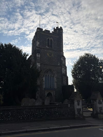The image displays St. Augustine's Church tower, located in Broxbourne, United Kingdom, viewed from a street-level vantage point. The central feature is a robust, square church tower, constructed from dark, irregular flint and dressed stone, rising against a partially cloudy sky. The tower culminates in a crenellated parapet, from which a vertical flagpole extends on the left and a weather vane with cardinal directions points right on the upper right corner. A circular clock face, featuring gold Roman numerals and hands indicating approximately 3:45, is positioned on the tower's third level from the top. Below this, a large, arched, multi-paned window is prominent, with smaller louvered arched windows on higher levels.
The tower is partially obscured by dark, dense trees flanking its sides. A low, dark stone wall, built from rough-hewn stones, defines the churchyard's boundary in the mid-ground. Behind this wall, several grey, weathered gravestones are visible. To the right, the wall integrates two square stone gate pillars with peaked caps, framing a dark, open metal gate leading into the churchyard. A black lamppost with a curved arm and antique-style lantern stands behind the right gate pillar.
In the foreground, an asphalt road with a yellow road line runs horizontally, separated from the stone wall by a concrete pavement. The upper portion of the image shows a sky filled with scattered white cumulus clouds, with some patches of blue sky visible. The lighting suggests the sun is positioned to create a partial silhouette of the church tower. No individuals or discernable actions are present in the scene.
