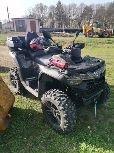 The image presents a black and camouflage all-terrain vehicle (ATV), with an "ONEAL" branded helmet placed on the seat, and a red extinguisher mounted on the front. The ATV is located in an outdoor setting. In the background, a yellow JCB tractor is visible. A small building with a light-colored facade is also present in the background. The ground is covered in green grass and other vegetation. The ATV is positioned on a partly bare patch of earth. To the left, there is a piece of yellow equipment. The picture was taken in Paisievo, Bulgaria.