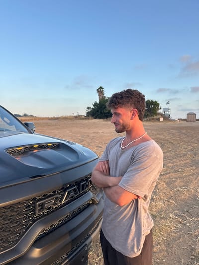 The image shows a young man standing next to a black Ram pickup truck. He is wearing a light gray t-shirt and black pants. The man is looking down and has his arms crossed. The truck is parked in a field of dirt and dry grass. The field appears to be a dirt parking lot. The sky is blue and there are a few trees in the distance. There is a palm tree in the distance on the right side of the image. The location is near Bnei Zion, Israel (32.226, 34.849). A white flag is flying in the distance. The image is taken from a low angle.