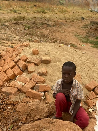 A child sits on a mound of sand surrounded by red bricks and large rocks. The child wears a plaid shirt and red pants, looking downwards with crossed arms. The ground surrounding the sand pile is brown and patchy with sparse vegetation on the left side. The overall scene appears to be an outdoor, possibly rural, environment.