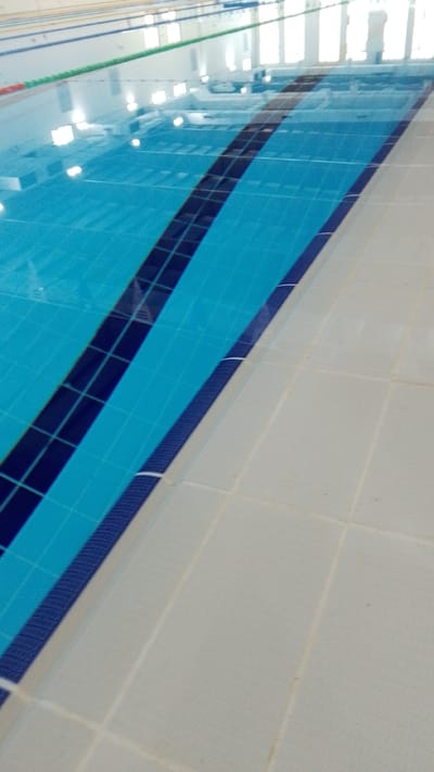 The image shows a tiled indoor swimming pool, likely in Umm Salal Ali, Qatar, with clear blue water and lane markers. The pool appears to be empty and unused, with sunlight reflecting off the water's surface and illuminating the ceiling. The surrounding area is also tiled, with a neutral color palette. There are no people or activities visible in the image, suggesting a quiet and serene environment. The overall impression is one of a well-maintained and modern aquatic facility.