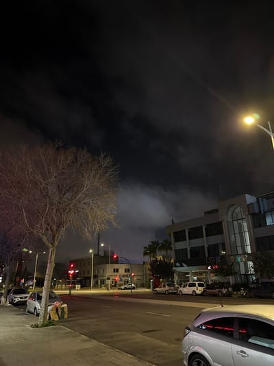 The image depicts a nighttime street scene in Polígon Industrial De Son Castello, Spain. A dark, heavily clouded sky covers the upper half of the frame, with brighter, illuminated sections of cloud visible in the mid-ground.
In the foreground, to the left, a large, bare deciduous tree stands on a sidewalk. The sidewalk, constructed of light-colored paving stones, runs along the left edge of the image. Adjacent to the tree, a silver hatchback vehicle is parked kerbside. Beside this vehicle, on the sidewalk, a stack of multiple cardboard boxes and other various items are accumulated.
The street itself appears to be a multi-lane asphalt road, partially visible, with multiple vehicles parked along both sides. Streetlights with yellow-orange illumination fixtures are visible on poles, casting light onto the scene. In the mid-ground, a series of buildings line both sides of the street. On the left, multi-story commercial buildings are visible, one with a partially discernible sign reading "QUILIC" or similar, and another with an illuminated red traffic signal showing a solid red light. A distinct plume or accumulation of white/grey smoke or mist rises from behind these buildings, spreading into the sky.
On the right side of the street, a multi-story building with a prominent arched window on its upper facade and dark-paned windows on lower levels is visible. Signs such as "LSI" and "MOTOS" are visible on its lower exterior. Several palm trees are situated near this building. No individuals are visible within the scene. The overall environment appears to be an urban or industrial area at night, under a cloudy sky with artificial illumination.