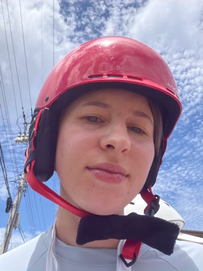 A close-up, selfie-style photograph shows a person from the chest up, wearing a red helmet with black ear pads and a red chin strap. The person has fair skin, light-colored eyes, and short brown hair visible around the helmet. The background is a bright blue sky with wispy white clouds. To the left, a utility pole with numerous wires is visible. The person appears to be outdoors. The image does not contain any specific geographical markers that would indicate it was taken in Seo-myeon, South Korea.