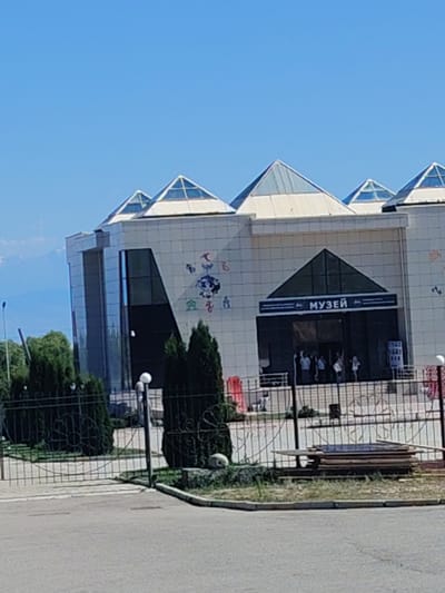 The image shows a modern museum building with a distinctive pyramid-shaped glass roof. The building has a light-colored facade with geometric window designs and is decorated with colorful murals. A sign above the entrance reads "МУЗЕЙ", which translates to "MUSEUM" in Russian. Several people are visible entering and exiting the building. The museum is surrounded by a black metal fence and lush green trees. In the background, distant mountains are visible against a clear blue sky. In the foreground, there is a paved road and a stack of wooden planks on the grass. The scene appears to be in Ornek, Kyrgyzstan, indicated by the prominent museum sign.
