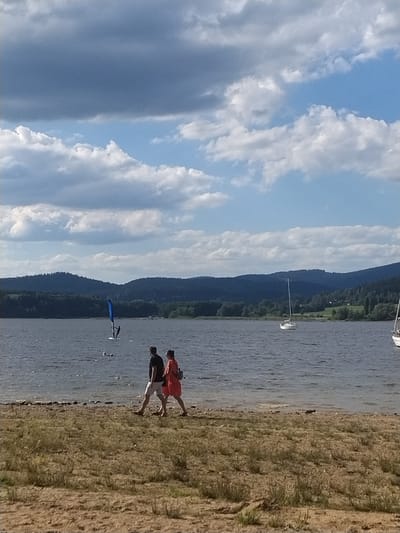 In Radslav, Czechia, on a body of water with dark blue, choppy waves, a person is windsurfing with a bright blue sail. In the distance, a white sailboat with a tall mast is visible on the water. The shoreline features a sandy beach with scattered dry grass and small plants. Two people are walking along the shoreline; the person on the left wears a black t-shirt and white shorts, while the person on the right is in a red dress. The background consists of lush green, tree-covered hills under a sky with dramatic, dark gray and white clouds interspersed with patches of bright blue.