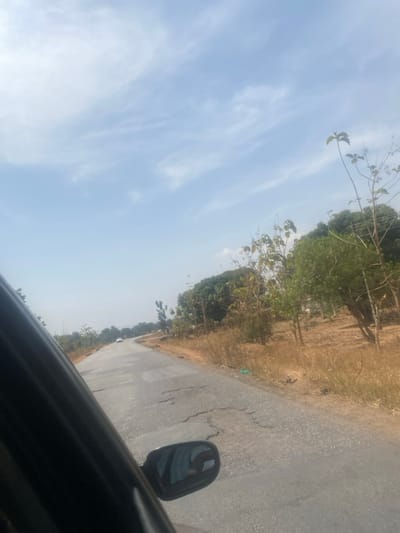 The scene depicts a cracked asphalt road in Aboro, Nigeria, viewed from the interior of a moving vehicle. The vehicle's dark-colored frame and a side mirror, reflecting a light blue object, occupy the lower left quadrant. The road surface exhibits extensive cracking and deterioration. In the distance, the road curves gently to the left, and a single white passenger vehicle is visible traveling ahead. The right roadside features dry, tan-colored grasses and scattered trees, some with green foliage and others appearing partially bare. Utility poles with overhead power lines run parallel to the road on the right. The sky is clear light blue with sparse, wispy white cloud formations, indicating daylight conditions. No individuals are explicitly visible in the scene.