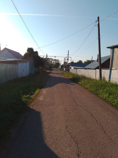 The image depicts a rural road in the city of Krasnokholmskiy, Russia, bathed in the warm light of either sunrise or sunset. The road, paved with asphalt and showing visible cracks, curves gently into the distance. On the left side of the road, a corrugated metal fence lines the property, adjacent to a low building with a weathered facade and a dark-colored roof. Further down the road, houses with metal roofs and wooden exteriors are visible, interspersed with trees. A prominent wooden utility pole stands on the right side of the road, supporting a network of overhead wires. The sky is a clear blue, with a faint white contrail stretching across it. Green grass and vegetation grow along the sides of the road and between properties, indicating a natural, uncultivated environment. The overall impression is one of a quiet, residential street in a Russian settlement.