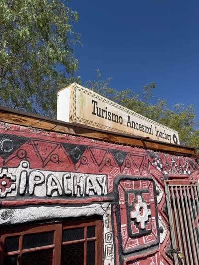 An outdoor, upward-angled view depicts the upper facade of a building under a clear, deep blue sky in Pica, Chile. Green trees with light foliage are visible in the upper background, partially obscuring the sky.
Mounted horizontally atop the building is a weathered, off-white rectangular sign framed by a dark border with a light patterned edge. It displays the text "Turismo Ancestral Ipachay" in dark, serif lettering. To the left of the text, a stylized yellow figure with outstretched limbs is depicted.
Below the sign, a rough wooden beam runs horizontally across the top of the building's painted facade. The facade is predominantly red and black, adorned with geometric patterns in white. The word "IPACHAY" is prominently rendered in raised white letters against a black background, framed by white borders.
To the lower left, a wooden-framed window with dark panes is visible beneath the "IPACHAY" text. To the right, a vertical barred grey metal gate or grille is partially visible, from which a brass-colored padlock is suspended.